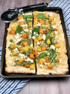 Butternut squash, arugula, ricotta & pumpkin seed topped gluten free pizza on a rectangular baking sheet.